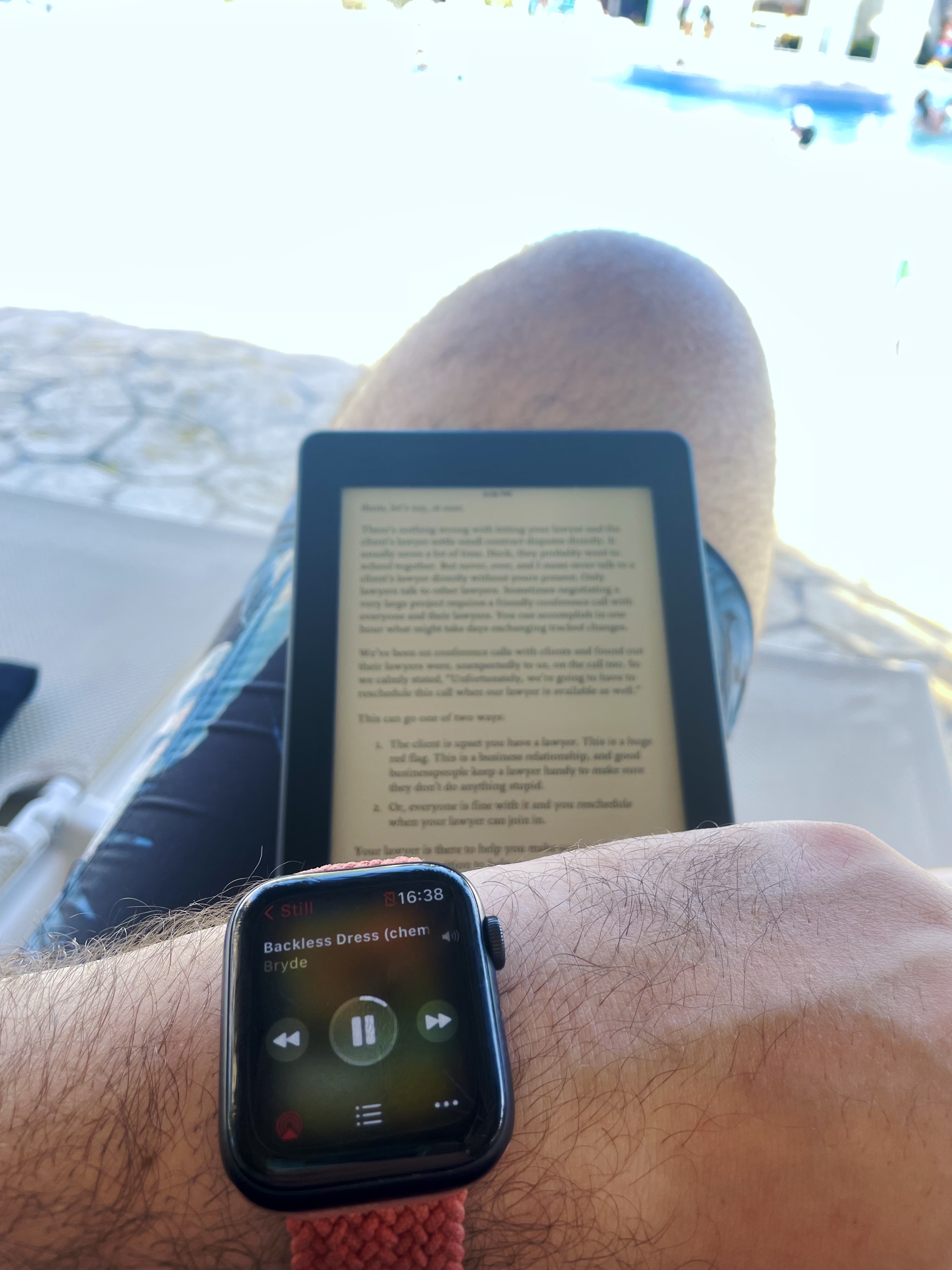Sitting on a sunbed looking over to the pool, Kindle on my lap, arm turned over to show Apple Watch now playing Backless Dress by Bryde.